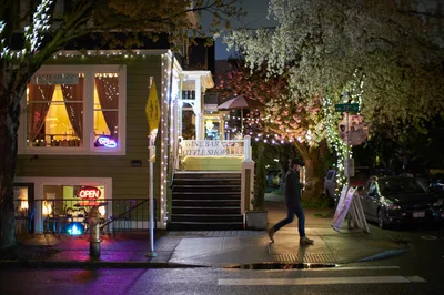 A solitary figure moves through the twilight streets of Portland's Nob Hill district, passing illuminated storefronts including a wine bar and bottle shop. Warm golden light spills from shop windows while delicate string lights trace the building's architectural lines, creating an intimate urban atmosphere. Spring blossoms on nearby trees add a seasonal softness to the hardscape, while purple neon signs cast colorful reflections on the wet pavement.