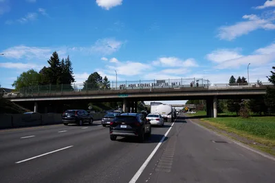 Activists display a protest banner reading 'No Work School Shopping May 1st' from a pedestrian overpass spanning Interstate 5 in North Portland's Humboldt neighborhood. The concrete bridge frames the banner against a brilliant blue spring sky dotted with white cumulus clouds, while morning traffic flows steadily beneath. Towering evergreens and mixed deciduous trees create a verdant backdrop, their fresh green foliage suggesting the renewal of spring organizing season.
