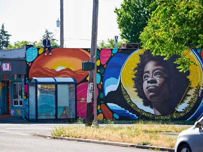 Travis Fields sits triumphantly atop a weathered rooftop, having just completed his vibrant mural honoring poet Amanda Gorman in Portland's Alberta district. The sprawling street art below bursts with warm sunset gradients, swirling organic shapes, and Gorman's portrait rendered with a golden halo against celestial blues. Dappled afternoon light filters through verdant tree canopy, casting the scene in the golden hour glow of creative accomplishment.