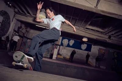 A skateboarder launches into weightless suspension above the concrete bowl at Commonwealth Skateboarding in Portland's Buckman neighborhood, arms spread wide against the industrial ceiling's exposed beams. The underground skate park's graffitied walls and scattered onlookers create a gritty urban cathedral where street culture thrives beneath fluorescent light. The moment captures pure athletic poetry—body extended in flight, board trailing below, while the venue's raw concrete and tagged surfaces frame this expression of underground artistry.
