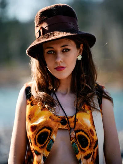 A young woman with flowing chestnut hair poses confidently along Oregon's Floodplain Trail, wearing a striking butterfly-patterned top and vintage brown fedora. Golden hour light creates dramatic rim lighting on her hair and hat while casting warm shadows across her face. Her bohemian styling includes flower earrings and layered necklaces, embodying free-spirited outdoor fashion against the softly blurred riverbed backdrop of Multnomah County's scenic landscape.