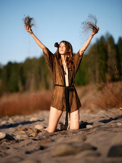 A young woman in an earth-toned robe kneels on the sandy riverbed along Oregon's Floodplain Trail, arms raised triumphantly as fine sand cascades from her open palms. The golden hour light bathes the scene in warm amber tones, while the blurred forest backdrop creates intimate depth. Her expression radiates pure joy and liberation, capturing a moment of spontaneous celebration amidst the natural riverside landscape of Multnomah County.
