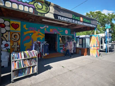 The vibrant Dime & Penny thrift store on Northeast 15th and Killingsworth displays its sustainable mission through bold murals and outdoor book displays. Afternoon sunlight illuminates the turquoise storefront adorned with colorful graffiti-style artwork featuring floating whales and abstract forms. A customer browses clothing racks beneath the covered entrance while rainbow-spined books create an inviting streetside library, embodying Portland's commitment to reuse and community exchange.