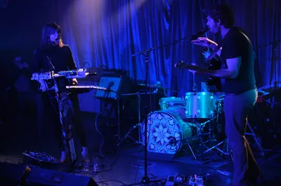 The indie duo Sun Blood Stories commands the intimate stage at Dante's in downtown Portland, bathed in ethereal blue and purple lighting that transforms the underground venue into a sonic sanctuary. A keyboardist in black manipulates synthesizers and electronic equipment on the left while a drummer anchors the rhythm from behind a kit adorned with geometric patterns, both silhouetted against flowing curtains that ripple with colored light. The atmospheric lighting creates dramatic shadows and highlights that emphasize the contemplative intensity of their performance at Rx Fest.