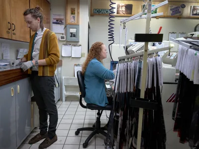 Two film lab technicians work in concentrated silence within the intimate confines of Blue Moon Camera & Machine in Portland's Saint Johns neighborhood. A woman with curly hair in a teal sweater sits at a processing station surrounded by hanging film strips, while her colleague in a mustard cardigan examines prints at the counter. The cramped workspace pulses with the quiet intensity of analog craftsmanship, fluorescent light casting clinical precision over rows of negatives and the accumulated ephemera of photographic tradition.