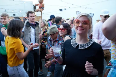 A blonde woman in a black dress with pearl collar and whimsical cat ear headband radiates joy while holding a cocktail at Your Sunday Best's season opener. The crowded rooftop gathering at White Owl Social Club buzzes with eclectic festival fashion, from vintage fur vests to yellow retro tops. Warm afternoon light bathes the diverse crowd of young adults mingling with drinks against the backdrop of Portland's urban skyline.