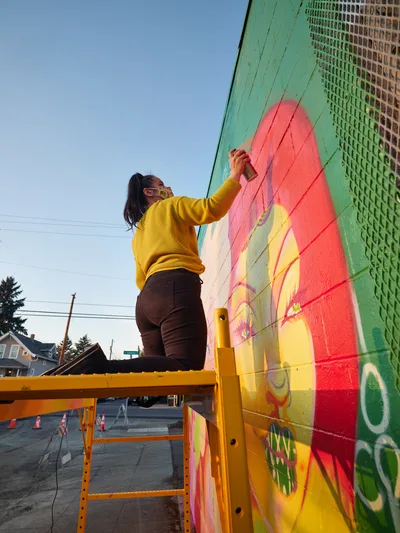 Multidisciplinary artist Villatose works from a yellow scissor lift, adding bold coral and yellow brushstrokes to her mural 'Jardín Cubana' featuring three female figures on Northeast Alberta Street in Portland's arts district. The late afternoon light bathes the scene as she meticulously applies paint to the mint green wall, her mustard sweater and dark pants creating a striking silhouette against the clear Oregon sky. Power lines and residential rooftops frame the background of this quintessential Portland neighborhood street art scene.