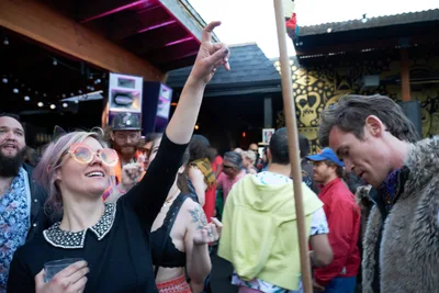 A jubilant woman with platinum blonde hair and round glasses throws her arm skyward in pure celebration, her face radiating joy as she dances among the crowd at White Owl Social Club's outdoor summer party. The warm afternoon light filters through the venue's industrial architecture, illuminating her black vintage-style dress and the diverse gathering of revelers around her. String lights and exposed beams frame the scene, while fellow dancers in colorful attire create a vibrant backdrop of community and connection.