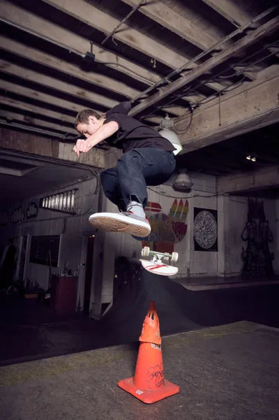 A young skateboarder launches into an aerial maneuver above an orange traffic cone at the dimly lit Commonwealth Skateboarding in Portland's Buckman neighborhood. The exposed wooden ceiling beams and graffitied concrete walls create an authentic underground atmosphere, while warm ambient lighting illuminates the skater mid-flight. Street art and skating culture merge in this intimate editorial moment captured in one of Portland's iconic skate shops.