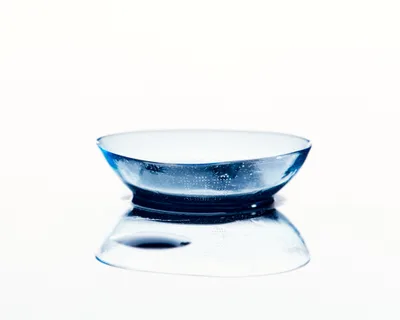A single contact lens rests delicately on a pristine reflective surface, its transparent form capturing light and creating a mesmerizing display of refraction. Tiny bubbles suspended within the hydrogel material sparkle like captured starlight, while the lens's gentle curve creates a perfect mirror reflection beneath. The minimalist composition against a pure white background transforms this everyday object into an elegant study of transparency and optical precision.