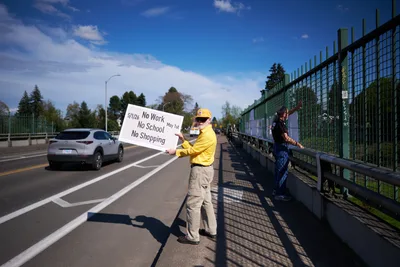 A lone activist in bright yellow jacket and cap stands on the North Skidmore Street overpass above Interstate 5 in North Portland, holding a handwritten protest sign calling for a May 1st General Strike. The demonstrator displays a banner reading "5/1/20 No Work No School No Shopping May 1st" while another person in dark clothing moves along the green metal fence. Afternoon sunlight casts sharp shadows across the concrete walkway as traffic flows beneath on the busy freeway, creating a powerful juxtaposition between grassroots activism and urban infrastructure.