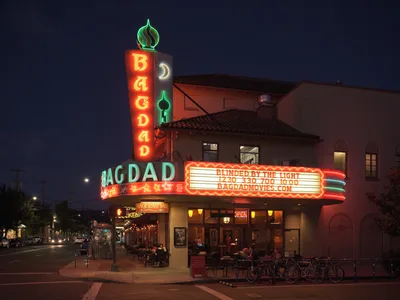 The historic Bagdad Theater & Pub radiates warmth against Portland's deepening twilight sky, its iconic vertical marquee blazing in red neon letters while the horizontal sign advertises showtimes. The Spanish Colonial Revival architecture frames the luminous entrance where patrons gather beneath the glowing canopy. Bicycles line the sidewalk and street lamps punctuate Southeast Hawthorne Boulevard, creating an intimate neighborhood cinema atmosphere in Richmond district.