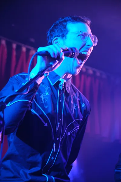 A vocalist from Roselit Bone commands the microphone under saturated blue and purple stage lighting at Dante's in Portland, Oregon. The performer's intense expression and wire-rimmed glasses catch the theatrical illumination as he leans into his delivery, creating a raw intimacy typical of the legendary venue's underground music scene. The moody lighting transforms the artist into a silhouette of electric energy against the club's shadowy interior. This captures the visceral connection between performer and audience that defines Portland's alternative music culture.