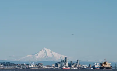 Mount Hood's snow-capped peak rises majestically beyond the industrial sprawl of the Port of Vancouver, Washington, as viewed from Kelley Point Park in Oregon. The crystalline morning light illuminates the mountain's glaciated slopes while cargo ships and port infrastructure create a striking juxtaposition of natural grandeur and maritime commerce. A small aircraft traces across the pale blue sky, adding scale to this panoramic vista where the Columbia River separates two states.