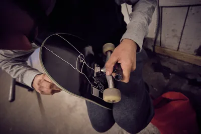 A skateboarder's weathered hands cradle a black skateboard deck adorned with intricate white line graphics, carefully adjusting the trucks and hardware in the warm, industrial atmosphere of Commonwealth Skateboarding in Portland's Buckman neighborhood. The intimate scene captures the ritualistic care between rider and board, with soft lighting illuminating the precise mechanical work. Red accents punctuate the shadowed workspace, while the concrete floor and utilitarian setting speak to the authentic culture of Southeast Portland's skateboarding community.