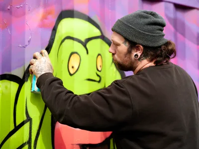 A bearded artist in a charcoal beanie and dark hoodie meticulously works on a bright lime green cartoon character against a vivid purple and pink backdrop. The intimate moment captures the focused concentration of street art creation, with the artist's tattooed hands delicately adding details to the whimsical figure's yellow eyes. The weathered urban wall provides texture and authenticity to this Portland neighborhood art scene.