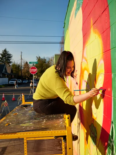 Multidisciplinary artist Vilatose works intently on her mural Jardín Cubana, depicting three female figures along Northeast Alberta Street in Portland's celebrated arts district. Golden hour light bathes the scene as she applies precise brushstrokes to the colorful wall, her yellow sweater echoing the warm tones of her artistic creation. The urban setting captures the creative energy of Portland's Alberta Arts District, where community murals transform neighborhood walls into canvases of expression.