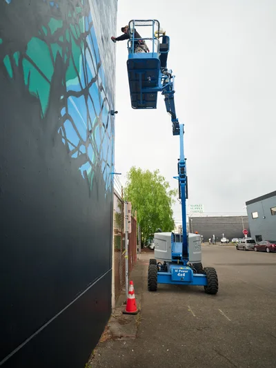 Artists Jeremy Nichols and Devin Finley work from the elevated platform of a blue cherry picker lift, applying vibrant green and blue paint to a large-scale mural on a concrete wall in Southeast Portland's Central Eastside district. The industrial setting features overcast skies and urban infrastructure, with the mechanical lift creating a dramatic vertical line against the sprawling street art composition. Safety cones mark the work zone while the artists concentrate on their collaborative public art project for the Portland Street Art Alliance.