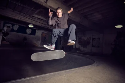 A skateboarder executes a dynamic flip trick above a concrete bowl at Commonwealth Skateboarding in Portland's Buckman neighborhood. The dramatic low-angle perspective captures the athlete suspended in mid-air, his board spinning beneath him as he extends both arms for balance. Moody artificial lighting illuminates the underground skate park's weathered concrete surfaces and graffitied walls, creating stark shadows that emphasize the intensity of the moment.