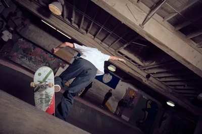 A skateboarder executes a dynamic aerial maneuver in the raw concrete basement of Commonwealth Skateboarding in Portland's Buckman neighborhood. The dramatic low-angle perspective captures the athlete mid-flight against exposed ceiling beams and weathered concrete pillars, while graffiti and street art provide colorful contrast to the industrial architecture. Moody lighting filters through the underground space, creating deep shadows that emphasize the gritty urban aesthetic of this Southeast Portland skate sanctuary.