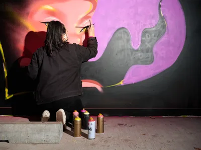 Multidisciplinary artist Vilatose delicately applies finishing touches to an eye detail within a large-scale mural featuring swirling purple and orange forms against a dark background. Working in the intimate confines of Clay Creative's parking garage in Portland's Hosford-Abernethy neighborhood, the artist crouches before their canvas with precise brush strokes while an array of spray paint cans waits nearby on the concrete floor. The dramatic interplay of warm coral highlights and cool violet shadows creates an otherworldly atmosphere in this underground creative sanctuary.