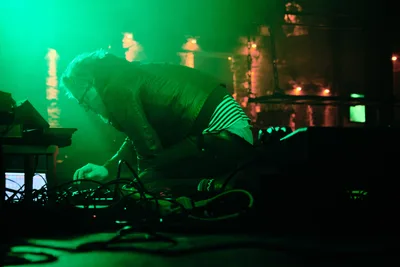 Electronic musician Eli Savage, performing as ellipsism, leans intimately over his synthesizer setup during a hypnotic set at RXFest 2019. Emerald stage lighting bathes the performer and his analog equipment in an otherworldly glow, while the iconic brick interior of Dante's provides a gritty backdrop to the ethereal soundscape. The image captures a moment of pure artistic immersion, as tangled cables and glowing displays create a technological altar of sound in Portland's legendary music venue.
