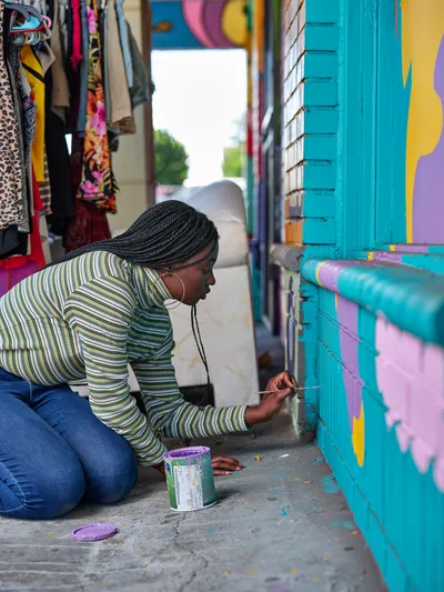 Naomi Likayi kneels on weathered concrete, carefully adding her signature to a vibrant turquoise and purple mural in Portland's Alberta neighborhood. Wearing a striped sweater and jeans, she works with focused concentration while colorful clothing hangs from a nearby storefront. The scene captures the intimate moment of artistic completion against the backdrop of Northeast Portland's creative commercial corridor.