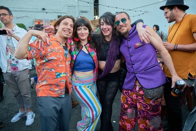 A jubilant group of five friends embraces in the golden afternoon light at White Owl Social Club's rooftop venue in Portland, Oregon. Their eclectic vintage-inspired outfits create a kaleidoscope of color against the urban backdrop, while string lights overhead hint at the festive atmosphere of the Your Sunday Best season opener. The genuine laughter and tight embrace capture the euphoric spirit of summer day parties, as revelers dressed in Hawaiian shirts, rainbow stripes, and purple blazers toast the beginning of another season of outdoor celebrations.