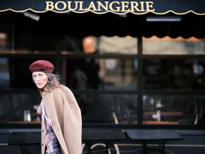 A young woman in a burgundy beret and camel coat glances back with an enigmatic expression as she passes a traditional French bakery storefront in Portland's Alberta Arts District. The warm golden lettering of 'BOULANGERIE' creates a romantic backdrop against the dark awning, while soft natural light illuminates her contemplative face. The composition captures a fleeting moment of urban European charm transplanted to the Pacific Northwest, with the subject's vintage-inspired styling echoing the old-world aesthetics of the bakery behind her.