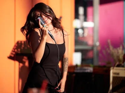 Aubrey Gordon leans into a vintage microphone with passionate intensity, her dark hair cascading as she performs at Portland Mercado in Southeast Portland. The warm amber and magenta stage lighting creates an intimate atmosphere in the cultural marketplace venue, while her tattooed arm and black ribbed top complement the moody concert setting. Her expression conveys pure musical joy as she connects with the audience through song.