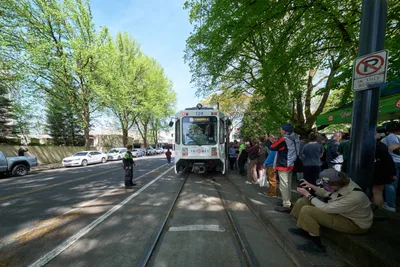 Enthusiasts gather along Northeast 11th Avenue in Portland's Lloyd District as TriMet's white Type 1 MAX train makes its ceremonial final run on April 18th, 2026. Dappled sunlight filters through mature street trees onto the crowd of transit fans and photographers positioned along the platform and trackside, creating a nostalgic atmosphere for this historic goodbye event. The scene captures the intersection of urban transit history and community celebration, with onlookers of all ages bearing witness to the end of an era in Portland's public transportation legacy.
