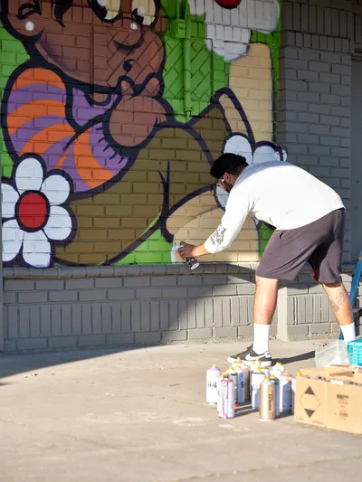 Muralist Heysus leans forward in concentrated effort, spray can in hand, adding intricate details to a colorful wall mural in Portland's Alberta Arts District. The artwork blooms across the brick surface with whimsical characters, bold florals, and a riot of greens, purples, and oranges. Scattered aerosol cans and a wooden crate of supplies rest on the sunlit sidewalk, evidence of the creative process unfolding in this corner of Northeast Portland where street art transforms urban landscapes.