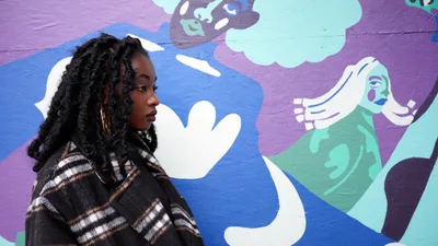 Artist Naomi Likayi sits in profile against her vibrant mural work at the boarded Portland World Trade Center, commissioned by the Portland Street Art Alliance. Her contemplative pose and striking locs create an intimate portrait against the dynamic backdrop of purple, blue, and mint green figures that seem to reach and interact across the wall. The interplay between the artist's earthtone plaid jacket and the bold street art creates a compelling juxtaposition of personal style and public expression.