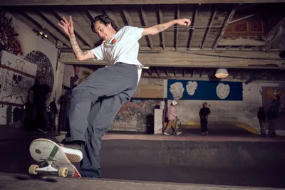 A tattooed skateboarder carves through Commonwealth Skateboarding's concrete bowl in Portland's Buckman neighborhood, his white shirt and grey pants contrasting against the graffiti-adorned walls. The underground skate park's raw industrial atmosphere comes alive with movement as spectators gather in the background beneath exposed ceiling beams. Warm tungsten lighting creates dramatic shadows across the weathered concrete surfaces, capturing the authentic energy of Portland's skateboarding culture.