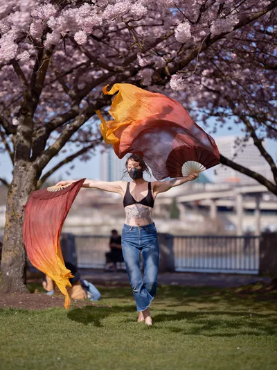A performer moves gracefully beneath the blooming cherry trees at Tom McCall Waterfront Park in Portland's Old Town district, her gradient silk fans creating flowing arcs of amber and coral against the pale pink sakura canopy. The masked dancer, dressed in casual jeans and bikini top with visible tattoos, embodies the intersection of contemporary street performance and traditional Asian dance forms. Urban architecture frames the distant background while dappled spring light filters through the delicate blossoms, capturing a moment where art, nature, and city life converge in Portland's historic Skidmore district.