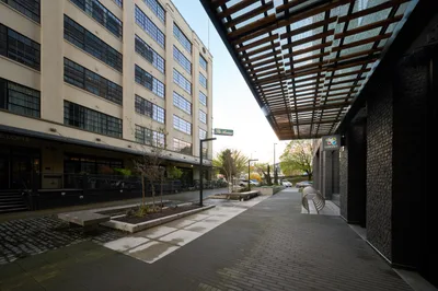 A contemporary urban courtyard in Portland's Pearl District captures the golden hour transition between converted industrial buildings. The geometric steel pergola overhead creates dramatic shadow patterns across wet brick pavers, while the restored warehouse facade with its signature grid windows anchors the composition. Warm interior lighting begins to emerge from ground-floor retail spaces as daylight fades, creating an atmospheric interplay between historic industrial architecture and modern urban renewal.