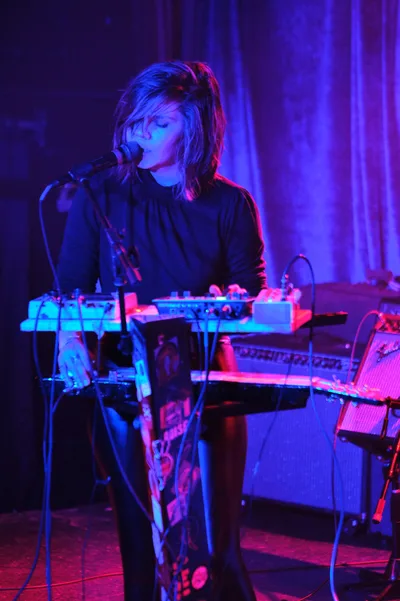A solo electronic musician performs at Dante's in downtown Portland, bathed in ethereal purple and magenta stage lighting. The artist, wearing dark clothing, leans into a microphone while surrounded by synthesizers and electronic equipment that glow with cyan and pink hues. The intimate venue's curtained backdrop creates a moody atmosphere as the performer becomes one with their synthesized soundscape during this Rx Fest showcase.