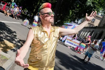 A charismatic parade participant in shimmering gold metallic attire commands attention with outstretched arms and rainbow-streaked hair beneath the dappled canopy of Portland's Pearl District trees. Soap bubbles drift through the sun-filtered air as colorful banners and enthusiastic crowds create a vibrant tapestry of celebration along Northwest 8th Avenue. The performer's theatrical pose and gleaming vest catch the filtered summer light, embodying the exuberant spirit of Pride festivities.