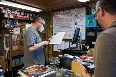 Inside the iconic Music Millennium record store on Portland's East Burnside Street during Record Store Day 2022, a masked employee in a gray t-shirt carefully reviews paperwork while a customer waits at the cluttered counter. The intimate shop buzzes with analog warmth, its wood-paneled walls plastered with vinyl records, concert posters, and a handwritten "New Releases" board listing upcoming albums. Fluorescent lighting casts an even glow over the transaction, capturing the enduring ritual of record store culture during this annual celebration of independent music retail in Oregon's music-loving city.