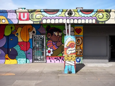 A kaleidoscopic street mural by artist Heysus 94 transforms the facade of Tuan's Auto Service in Portland's Alberta Arts District. The exuberant artwork features a menacing green skull with bared white teeth dominating the upper portion, while playful cartoon characters, abstract shapes, and bold typography cascade across the brick wall below. Bright afternoon light illuminates the saturated colors—electric blues, cherry reds, lime greens, and sunny yellows—creating a striking contrast against the weathered concrete sidewalk and clear blue sky.