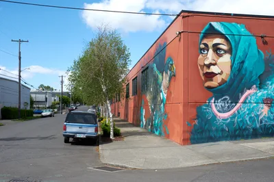 A monumental mural by artists Suhaib Attar and Ernesto Maranje transforms a rust-colored industrial building on Southeast Washington Street, depicting a woman with striking turquoise hair and penetrating gaze. The portrait emerges from flowing aquamarine brushstrokes against the building's weathered facade, while power lines and a leafy birch tree frame the scene under Oregon's characteristic cloudy sky. Part of Portland Street Art Alliance's community engagement initiative, this aptART-commissioned work brings vibrant artistic expression to the Central Eastside's industrial landscape.