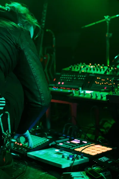 Electronic musician Eli Savage, performing as ellipsism, commands a complex array of synthesizers and electronic equipment bathed in ethereal green stage lighting at Portland's legendary Dante's venue. The intimate underground atmosphere captures the artist mid-performance, leather jacket and flowing hair silhouetted against the glowing constellation of buttons, knobs, and LED displays. Multiple levels of electronic instruments create a technological landscape while warm amber and cool green lights paint the scene in cyberpunk hues.