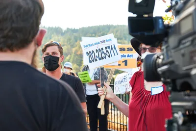 Michael Gaskill, wearing a black face mask, faces a television camera during a climate change protest outside Representative Kurt Schrader's office in Oregon City, Oregon. A demonstrator in period dress holds a protest sign displaying a phone number, while autumn-touched hills form a scenic backdrop. The scene captures the intersection of grassroots activism and media coverage, with bright afternoon light illuminating the passionate exchange between citizen and journalist.