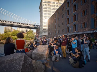 A vibrant gathering of photographers and onlookers assembles along the cobblestone waterfront beneath the Manhattan Bridge in Brooklyn's DUMBO neighborhood. The late afternoon light bathes the historic brick warehouses and modern high-rises in warm golden tones, while dozens of camera-wielding enthusiasts focus their lenses on subjects positioned among weathered granite boulders. The iconic suspension bridge cables stretch dramatically overhead, framing this quintessential New York photography scene where industrial heritage meets contemporary urban culture.