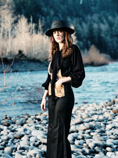 A contemplative woman in flowing black attire and wide-brimmed hat stands among the weathered river stones of Oregon's Floodplain Trail. Golden hour light illuminates her auburn hair and creates a dreamy atmosphere against the backdrop of dormant winter grasses and the flowing waters of Multnomah County's riparian landscape. The scene captures a moment of quiet reflection in the Pacific Northwest wilderness.
