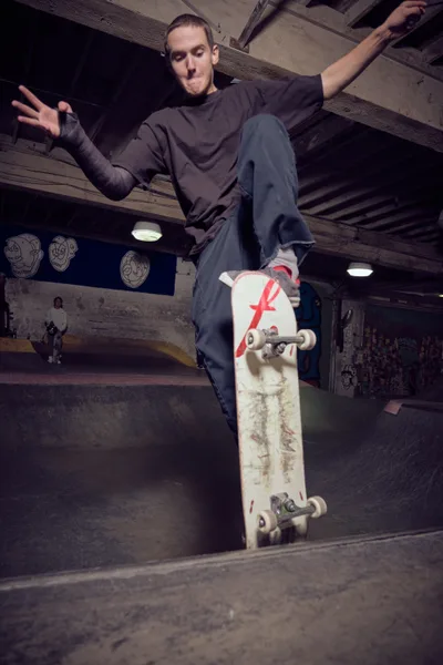 A skateboarder suspended mid-air above a concrete bowl at Commonwealth Skateboarding in Portland's Buckman neighborhood, his weathered deck marked with a distinctive red 'F' logo. The dramatic low-angle perspective captures the moment of weightless transition, arms outstretched for balance against the industrial backdrop of exposed ceiling beams and graffitied walls. Harsh artificial lighting creates stark contrasts in this subterranean skate sanctuary.