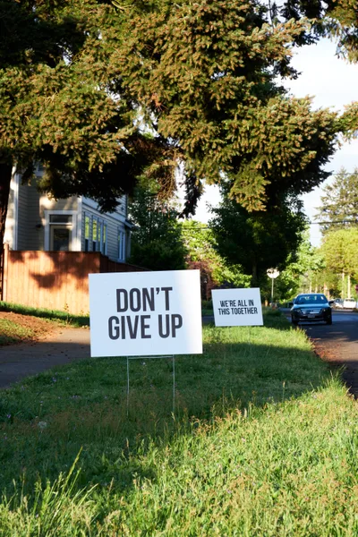 Two white lawn signs bearing messages of encouragement—"DON'T GIVE UP" and "WE'RE ALL IN THIS TOGETHER"—stand sentinel along Northeast Going Street in Portland's Beaumont-Wilshire neighborhood. Golden afternoon light filters through the canopy of mature evergreens, casting dappled shadows across the verdant grass while residential homes and a parked sedan create an intimate tableau of community solidarity during uncertain times.