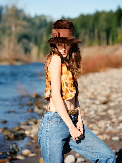 A young woman poses confidently along the rocky riverbed of Oregon's Floodplain Trail, her brown felt hat casting dramatic shadows across her face. She wears a vibrant tie-dye tank top in sunset hues paired with high-waisted denim, creating a striking contrast against the serene blue water and distant forest. The warm golden hour light bathes the scene, highlighting the natural textures of scattered stones and creating an intimate, contemplative mood.