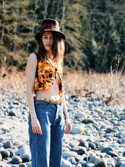A young woman stands contemplatively among the sun-bleached stones of a dried riverbed along Oregon's Floodplain Trail. Golden afternoon light illuminates her vintage-inspired ensemble of high-waisted denim, a tie-dye crop top, and weathered leather hat, while autumn grasses and bare cottonwoods create a textured backdrop. Her direct gaze and relaxed stance suggest a moment of peaceful solitude in this riparian landscape near the Columbia River Gorge.