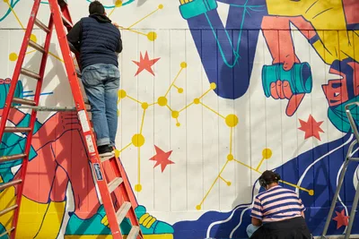 Two muralists work at different heights on a vibrant public art installation titled 'Let's Talk' at Open Signal in Portland's historic Eliot neighborhood. One artist climbs a red step ladder while another crouches near the base, both contributing to a dynamic composition featuring bold geometric figures, golden constellation-like networks, and scattered red stars against white concrete block walls. The collaborative scene captures the methodical process of large-scale community art creation in Northeast Portland's cultural corridor.